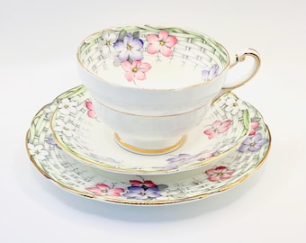 May include: A vintage teacup, saucer, and side plate set. The white porcelain features a delicate floral design with pink and purple flowers, a woven basket pattern, and gold trim. The teacup has a curved handle.