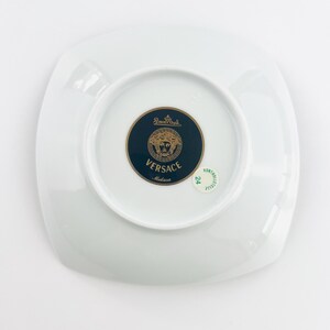 May include: A white, square-shaped plate with a glossy finish. The center features a dark blue and gold medallion with the Versace logo and the words "Versace Medusa". The plate is a piece of luxury tableware.