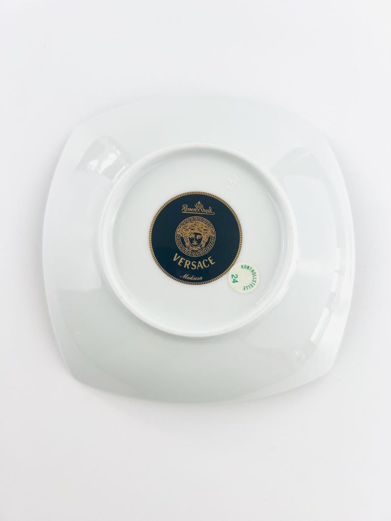 May include: A white, square-shaped plate with a glossy finish. The center features a dark blue and gold medallion with the Versace logo and the words "Versace Medusa". The plate is a piece of luxury tableware.