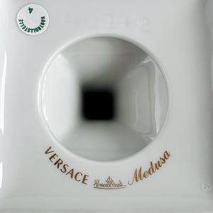 May include: A white porcelain object with a central circular opening and a square base. The words "VERSACE Medusa" are printed in gold, along with the Rosenthal logo. A small green sticker is visible.