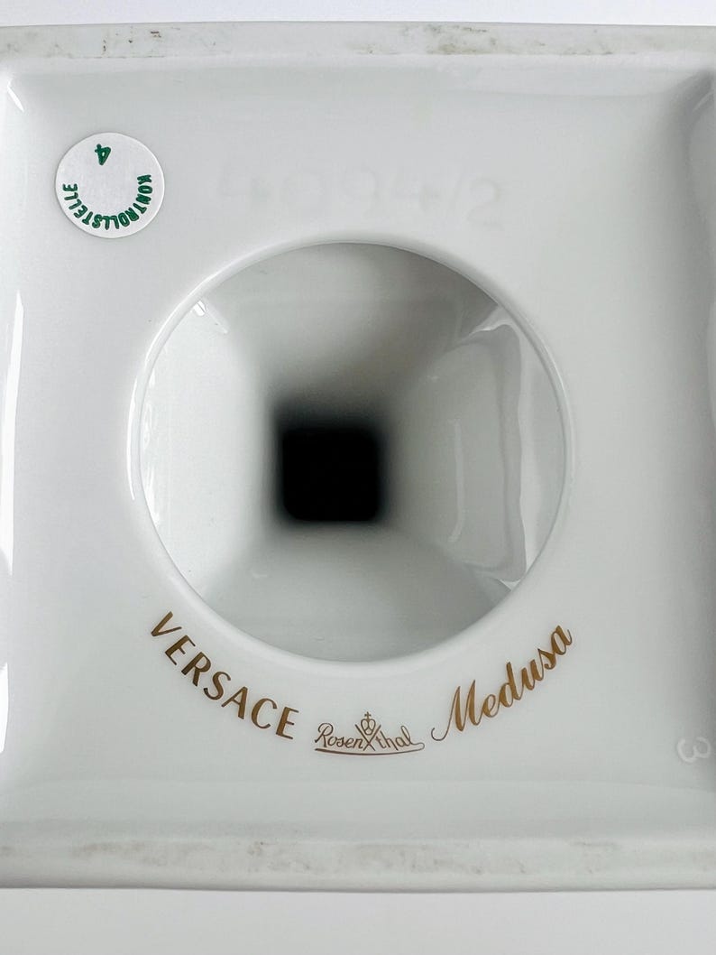 May include: A white porcelain object with a central circular opening and a square base. The words "VERSACE Medusa" are printed in gold, along with the Rosenthal logo. A small green sticker is visible.