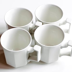 May include: Four white ceramic mugs with geometric sides and square handles are arranged on a white surface. The mugs are empty and have a simple, modern design. The mugs are suitable for coffee, tea, or other beverages.