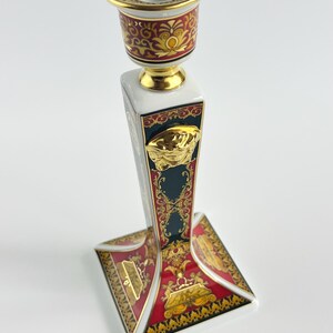 May include: A decorative ceramic candlestick holder with a white, red, and gold design. The candlestick features a square base and a cylindrical top, with ornate patterns and a gold Medusa head emblem.