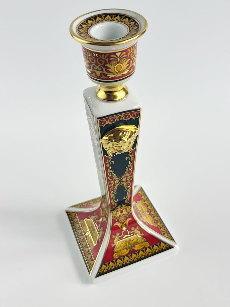 May include: A decorative ceramic candlestick holder with a white, red, and gold design. The candlestick features a square base and a cylindrical top, with ornate patterns and a gold Medusa head emblem.