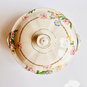 May include: A cream-coloured vintage serving dish with a lid, decorated with a floral pattern in pink, blue, and yellow. The dish has a scalloped edge and a central handle. The rim is accented with a gold trim. The design is reminiscent of classic tableware.