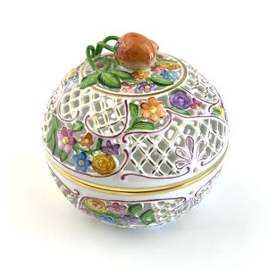 Può includere: Un portagioie rotondo in porcellana bianca con un motivo a griglia e accenti floreali colorati. Il coperchio presenta un pomello a forma di frutto marrone con foglie verdi. I bordi della scatola sono rifiniti con bordi dorati.