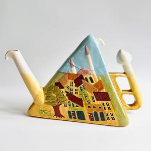 Puede incluir: Una tetera de cerámica con un diseño triangular único, que presenta una escena de pueblo pintada. La tetera tiene un fondo de cielo azul claro, con detalles en marrón, verde y amarillo. El pico y el asa son amarillos y blancos.