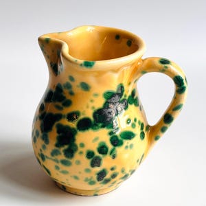 Puede incluir: Un pequeño jarro de cerámica amarilla con asa y pico vertedor. El jarro está decorado con manchas moteadas de color verde oscuro. El asa es un lazo redondeado y el pico vertedor está ligeramente curvado. El jarro probablemente se utiliza para servir líquidos.