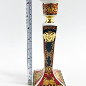 May include: A decorative ceramic candlestick holder with a gold, red, and black design. The candlestick is approximately 8 inches tall. The base features a red and gold pattern, with a gold Medusa head emblem.