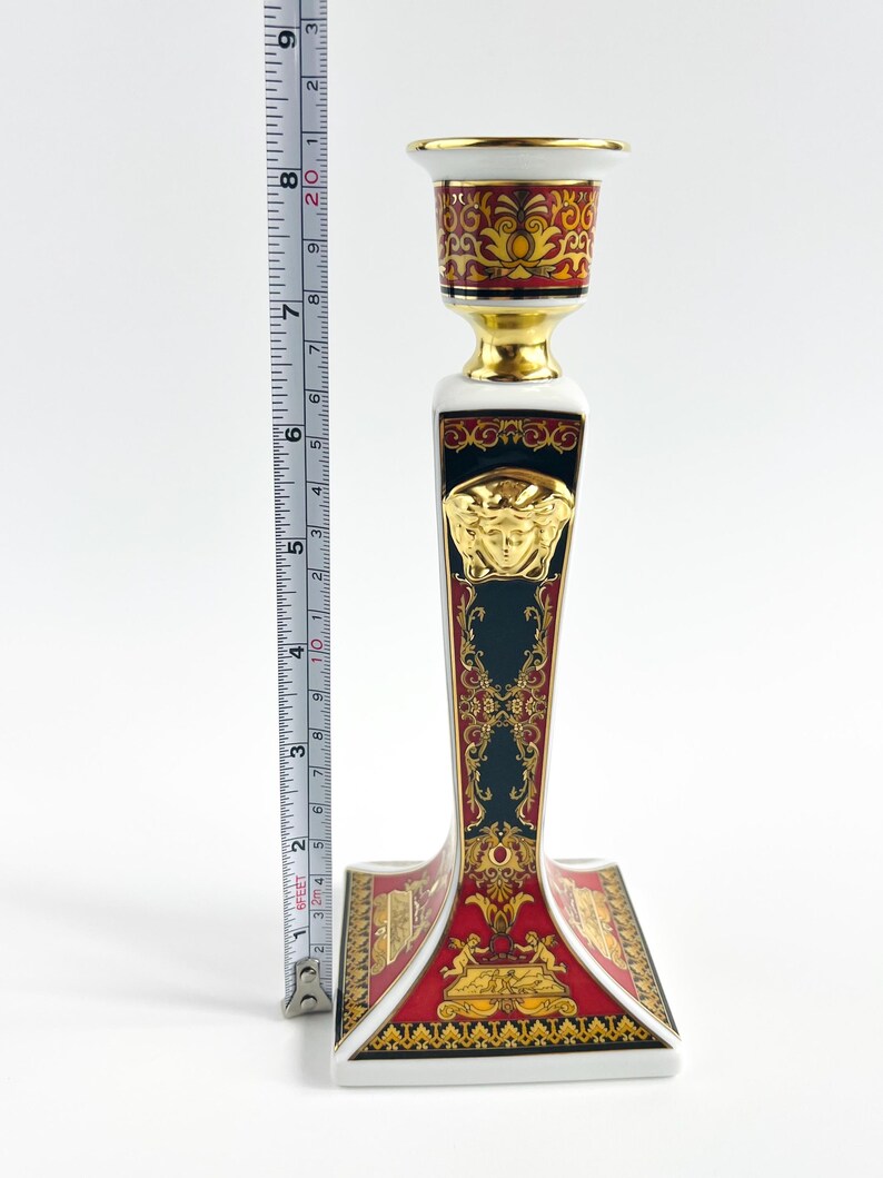 May include: A decorative ceramic candlestick holder with a gold, red, and black design. The candlestick is approximately 8 inches tall. The base features a red and gold pattern, with a gold Medusa head emblem.