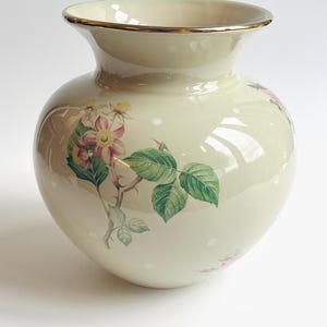 May include: Cream-colored ceramic vase with a gold rim and floral design. The vase features a rounded body and a flared opening. The floral design includes pink and yellow flowers with green leaves and stems. White polka dots are scattered across the surface.