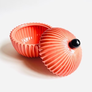 May include: Coral-colored ceramic trinket box with a ridged, shell-like design. The lid features a black, spherical knob. The box is open, revealing the interior. The box is on a white background.