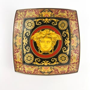 May include: Square decorative plate with a black, red, and gold ornate design. The center features a gold Medusa head on a black background, surrounded by a red border. The plate's edges have a detailed pattern.