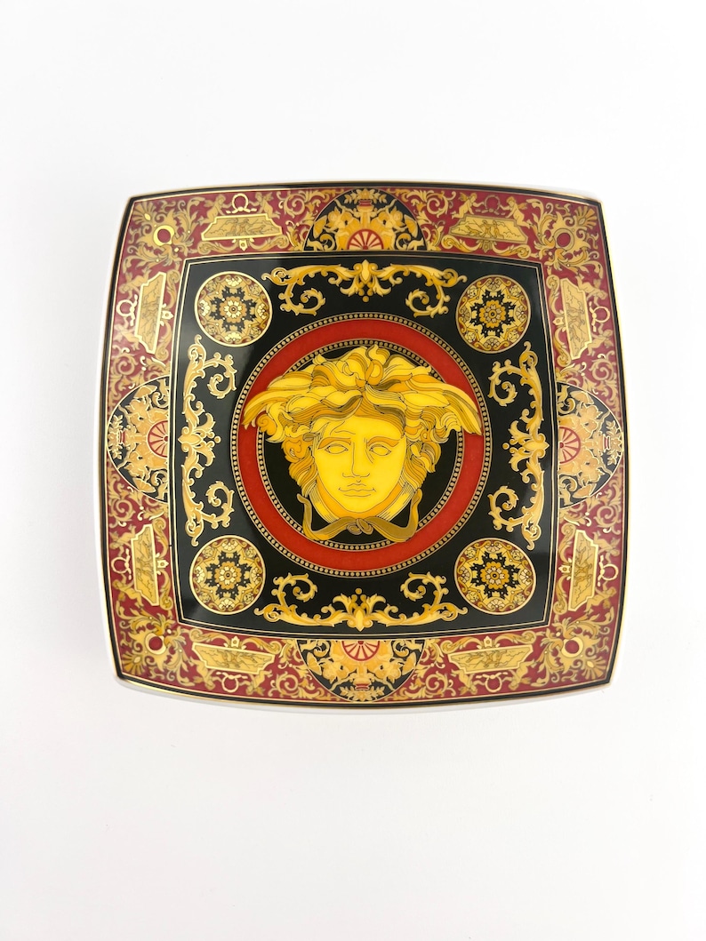 May include: Square decorative plate with a black, red, and gold ornate design. The center features a gold Medusa head on a black background, surrounded by a red border. The plate's edges have a detailed pattern.