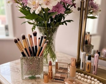 May include: A collection of makeup brushes in a square glittery holder, a lipstick, an eyeshadow palette, a foundation bottle, a compact powder, and perfume atomizers arranged near a glass vase filled with pink and white flowers.