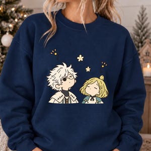 Op de afbeelding: Een marineblauw sweatshirt met ronde hals voorzien van een grafische illustratie van twee stripfiguren met wit en blond haar, omringd door kleine gele bloemen en zwevende stippen, gedrukt op het midden van de borst.