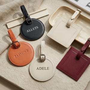 Peut inclure: Plusieurs étiquettes de bagages en cuir de différentes formes et couleurs. Les étiquettes comprennent des modèles ronds et rectangulaires en noir, orange, crème et bordeaux. Chaque étiquette comporte une sangle avec une boucle et un texte personnalisé, comme "ALLETS" et "JASMINE".