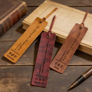 Könnte beinhalten: Drei Leder-Lesezeichen in Braun- und Burgundertönen, jedes mit einer Schlaufe und einem Band. Die Lesezeichen weisen eingravierte Zitate, Initialen und Daten auf. Sie sind auf einer Holzoberfläche in der Nähe eines Buches und eines Stifts angeordnet.