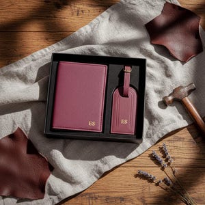 May include: A burgundy leather travel set, including a passport holder and luggage tag, presented in a black gift box. The items are embossed with the initials "ES" in gold. The set is displayed on a wooden surface with a linen cloth.