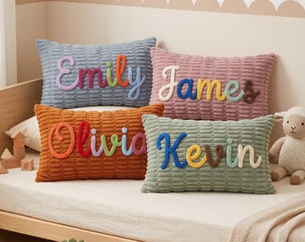 Puede incluir: Cuatro cojines rectangulares con una superficie de tela texturizada y acanalada. Cada cojín presenta un nombre personalizado bordado con letras de hilo coloridas y en relieve. Los nombres mostrados son Emily, James, Olivia y Kevin, en letra cursiva.