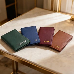 May include: Four personalized leather wallets in green, blue, burgundy, and brown, each with initials and symbols. The green wallet says "I LOVE U". The wallets are arranged on a marble-like surface.