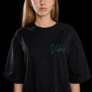Peut inclure: T-shirt noir à manches courtes. Le mot "Vibes" est écrit en vert en écriture cursive sur le devant. Le t-shirt a une coupe décontractée et est fait d'une matière douce. Le fond est noir.