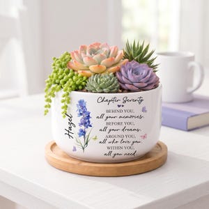May include: A white ceramic planter on a round wooden base, filled with various green, pink, and purple succulents. The planter features the text "Chapter Seventy" and a quote about memories, dreams, and love, alongside a blue flower and butterflies.