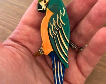 May include: A hand-painted wooden parrot figurine. The parrot features a teal head and wings, an orange chest, and a blue tail. The bird's beak is black, and the design includes white and gold accents.