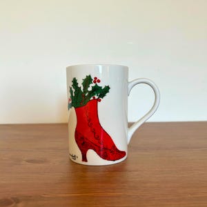 May include: White ceramic mug featuring a red high-heeled boot design filled with green holly and red berries. The mug has a curved handle and is suitable for hot beverages. The design is festive and seasonal.