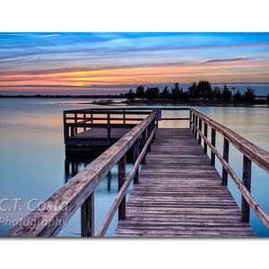 May include: A wooden dock extends out over a calm lake at sunset. The sky is a mix of blue, pink, and orange. The dock has a wooden railing and leads to a small wooden platform.