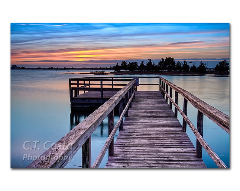 May include: A wooden dock extends out over a calm lake at sunset. The sky is a mix of blue, pink, and orange. The dock has a wooden railing and leads to a small wooden platform.