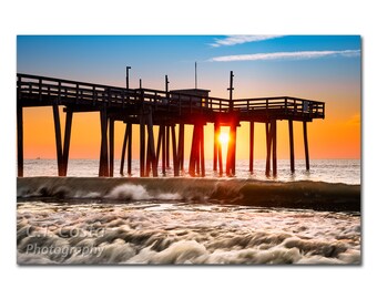 May include: A wooden pier extends over a body of water with a bright orange sunrise in the background. The sun is shining through the pier's beams, creating a starburst effect. The water is choppy and white-capped.