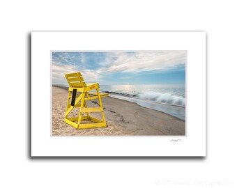 May include: A yellow lifeguard chair sits on a sandy beach with a blue sky and ocean in the background.