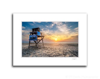 May include: A wooden lifeguard stand with blue and white signage on a sandy beach at sunrise. The sun is rising over the ocean in the background.