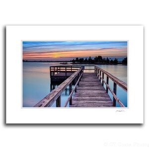 May include: A wooden jetty stretches out into a tranquil lake at sunset. The sky is a blend of pink, orange, and blue, with clouds mirrored in the water. The jetty is constructed from weathered wood with handrails on both sides.