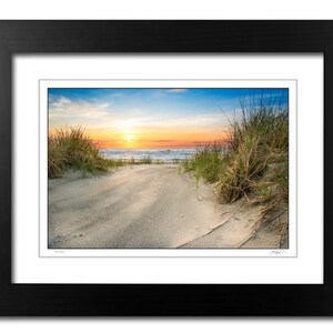 May include: A scenic photograph of a sandy beach path leading to the ocean. The path is lined with tall green grasses and the ocean is in the background with a bright orange and blue sunset sky.