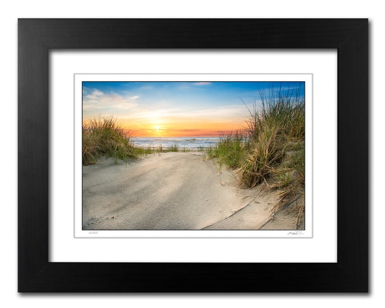 May include: A scenic photograph of a sandy beach path leading to the ocean. The path is lined with tall green grasses and the ocean is in the background with a bright orange and blue sunset sky.