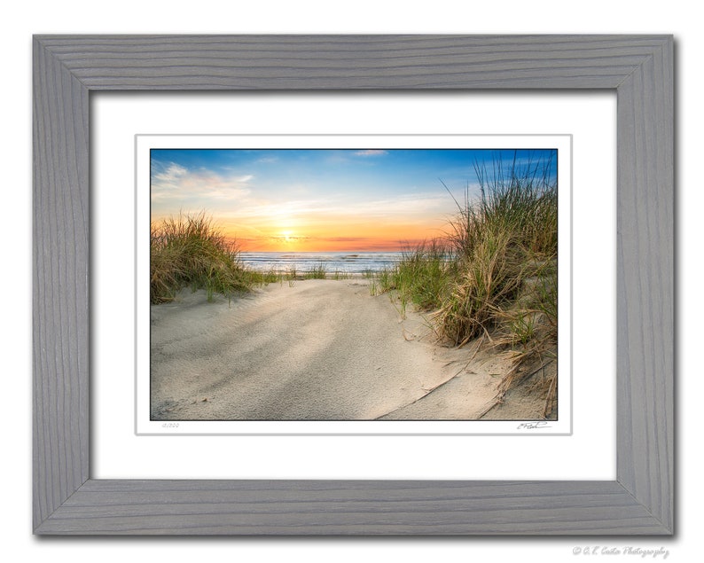 May include: A photograph of a beach scene with a path leading through the sand dunes to the ocean. The sky is a vibrant blue with a bright orange sunset in the distance. Tall grasses line the path.