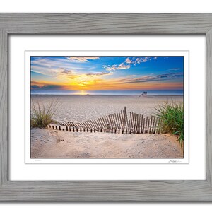 May include: A photograph of a beach scene with a wooden fence in the foreground. The beach is sandy and the ocean is in the background. The sky is blue with clouds and the sun is setting. The image is framed in a light gray frame.
