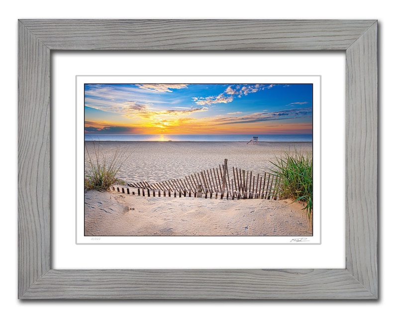 May include: A photograph of a beach scene with a wooden fence in the foreground. The beach is sandy and the ocean is in the background. The sky is blue with clouds and the sun is setting. The image is framed in a light gray frame.