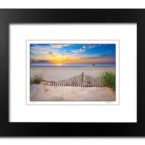 May include: A photograph of a beach scene at sunrise with a wooden fence in the foreground. The sky is a vibrant blue with pink and orange clouds. The beach is sandy and there is a lifeguard stand in the distance.
