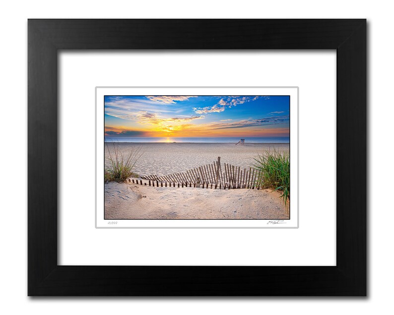 May include: A photograph of a beach scene at sunrise with a wooden fence in the foreground. The sky is a vibrant blue with pink and orange clouds. The beach is sandy and there is a lifeguard stand in the distance.