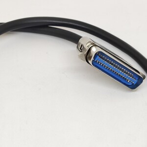 K&ouml;nnte beinhalten: Ein schwarzes Kabel mit einem silbernen Stecker. Der Stecker hat einen blauen Innenraum mit mehreren Pins. Das Kabel ist gebogen und der Hintergrund ist wei&szlig;.