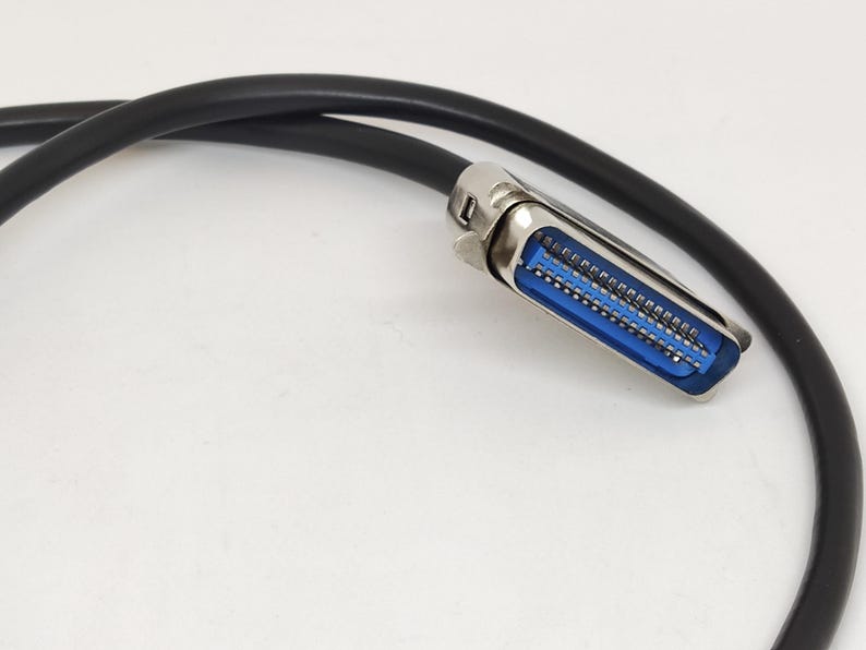 K&ouml;nnte beinhalten: Ein schwarzes Kabel mit einem silbernen Stecker. Der Stecker hat einen blauen Innenraum mit mehreren Pins. Das Kabel ist gebogen und der Hintergrund ist wei&szlig;.