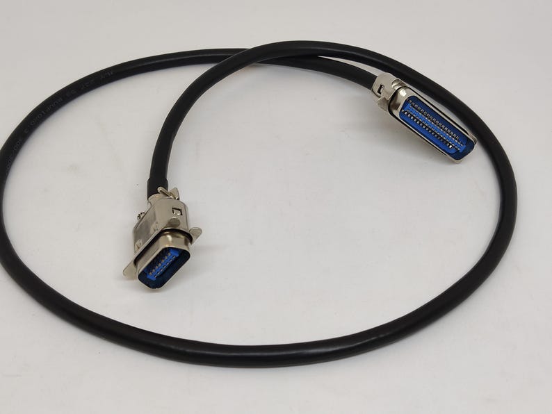 K&ouml;nnte beinhalten: Ein schwarzes Kabel mit zwei silbernen Steckern, jeweils mit blauem Innenraum. Das Kabel ist geschlungen, und die Stecker befinden sich an jedem Ende. Die Stecker haben ein D-f&ouml;rmiges Design.