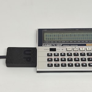 Puede incluir: Una computadora personal Casio FX-880P con un dispositivo rectangular negro adjunto. La calculadora tiene una pantalla que muestra números y texto. El dispositivo adjunto a la calculadora tiene la palabra "UP" impresa.
