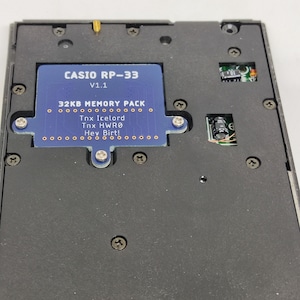May include: A close-up of a black electronic device with a blue circuit board labeled "CASIO RP-33 V1.1 32KB MEMORY PACK." The text "Tnx Icelord, Tnx HWRO, Hey Birt!" is also visible. The device is secured with multiple screws.