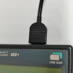 Puede incluir: Primer plano de un cable negro conectado a una calculadora Hewlett Packard 48G+. La calculadora es de color gris oscuro con una pantalla verde y el texto "128K RAM". El cable está conectado en la parte superior de la calculadora.