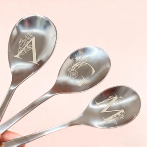 Puede incluir: Tres cucharas de acero inoxidable con diseños florales grabados y las letras A, G y M. Las cucharas tienen una superficie brillante y reflectante, sobre un fondo rosa claro. Las letras están en una fuente serif.