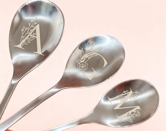 Può includere: Tre cucchiai in acciaio inossidabile con disegni floreali incisi e le lettere A, G e M. I cucchiai hanno una superficie lucida e riflettente, su uno sfondo rosa tenue. Le lettere sono in un carattere serif.